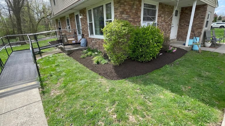 Front view of brick ranch house with manicured lawn, mulched landscaping, wheelchair ramp, and metal railings on a sunny day