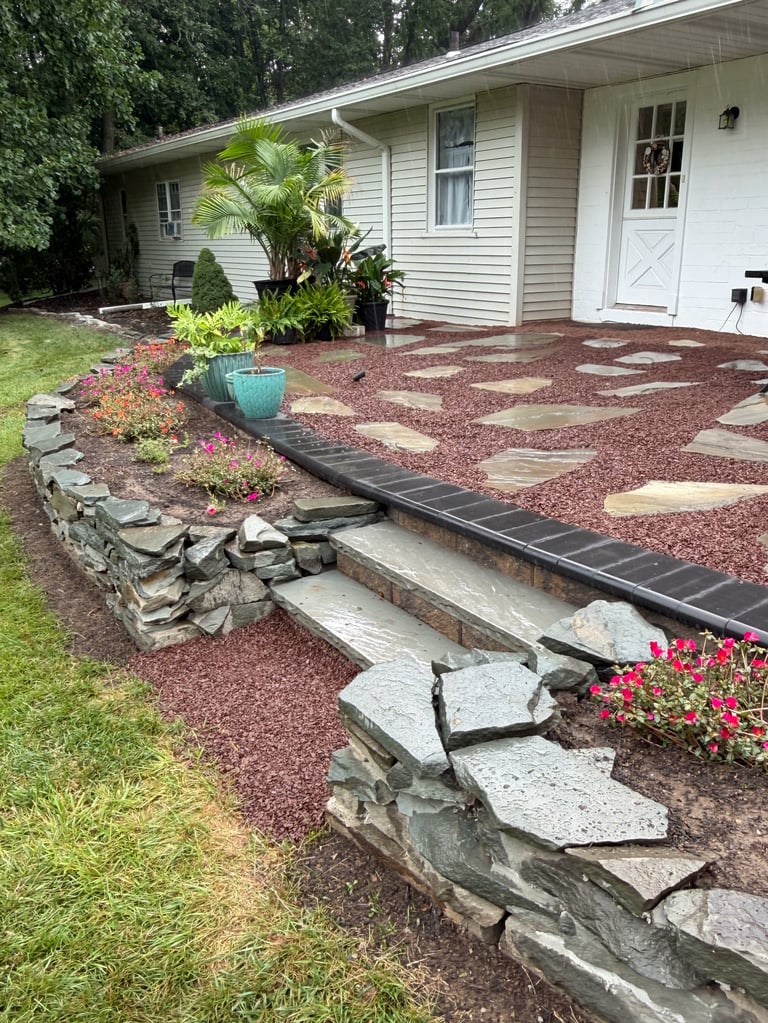 White single-story house with stone steps leading to landscaped front yard featuring red mulch, stepping stones, potted plants, and pink flowers
